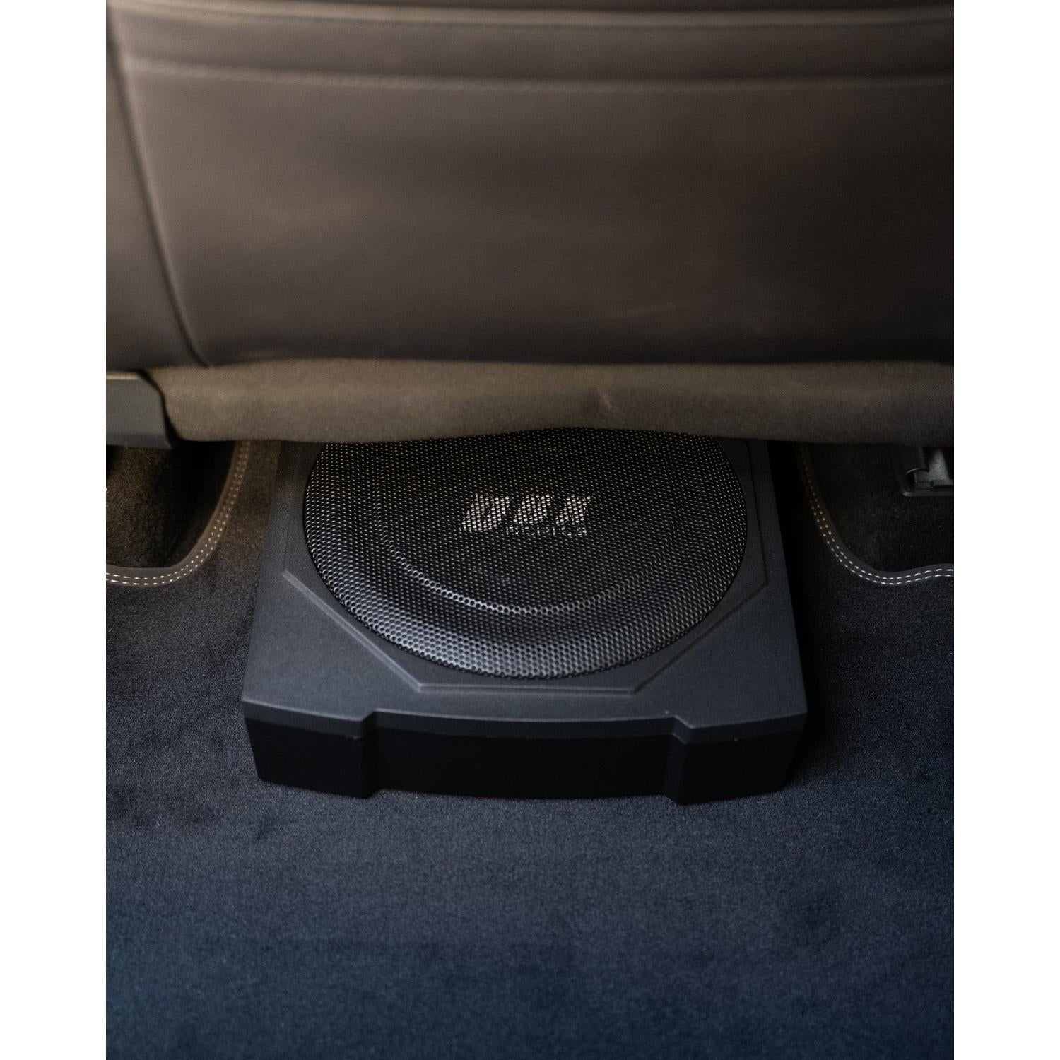 El Edge Car Audio EDBX10A-E0, un subwoofer negro de 10 Zoll con el logotipo MBX, se sitúa bajo un asiento de coche con diseño ultraflache entre la moqueta oscura y las costuras visibles del asiento.