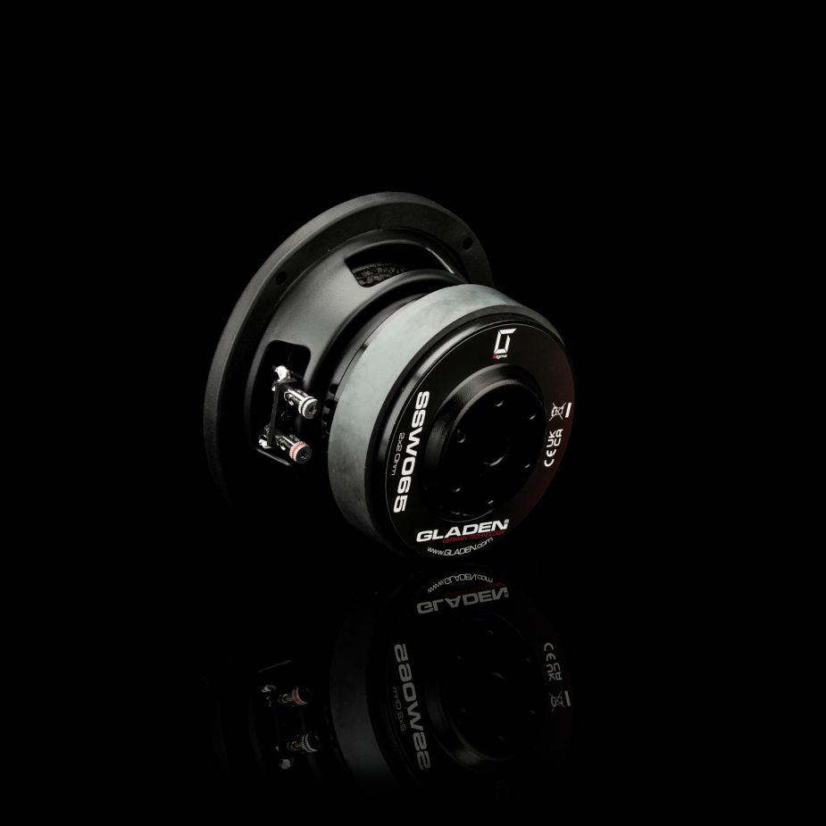 El Gladen Sigma SSW065, un subwoofer negro de 5 pulgadas con el centro y los terminales plateados, perfecto para su instalación en coches compactos, se muestra sobre una superficie negra reflectante.