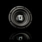 Un subwoofer Gladen Sigma SSW08 negro con un borde metálico aparece situado en el centro de una superficie negra reflectante sobre un fondo oscuro.