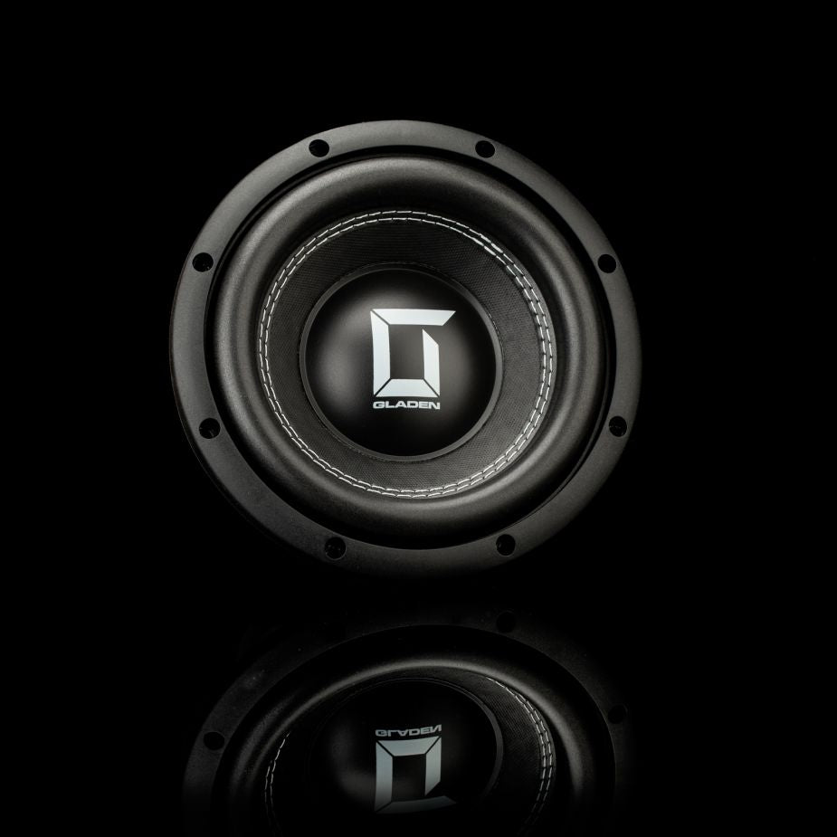 Un subwoofer Gladen Sigma SSW08 negro con un borde metálico aparece situado en el centro de una superficie negra reflectante sobre un fondo oscuro.