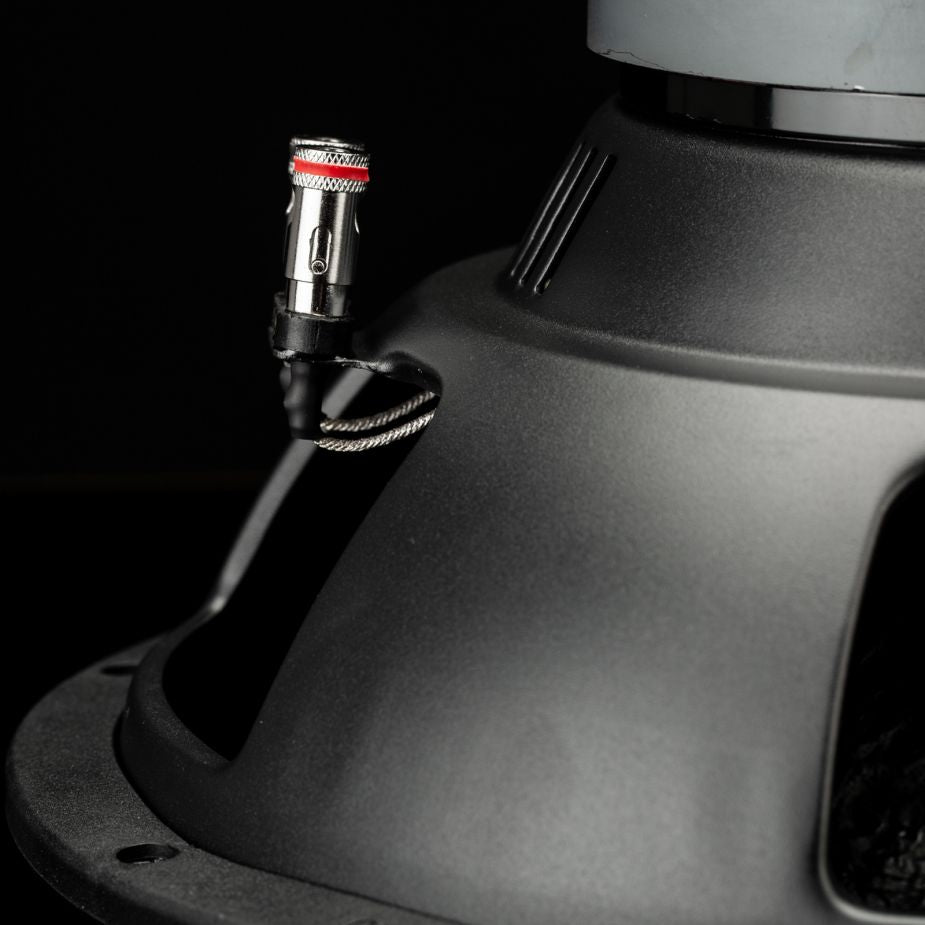 Primer plano de un terminal del subwoofer Gladen Sigma SSW12 con un conector de cable negro con una banda roja, sobre un fondo oscuro.