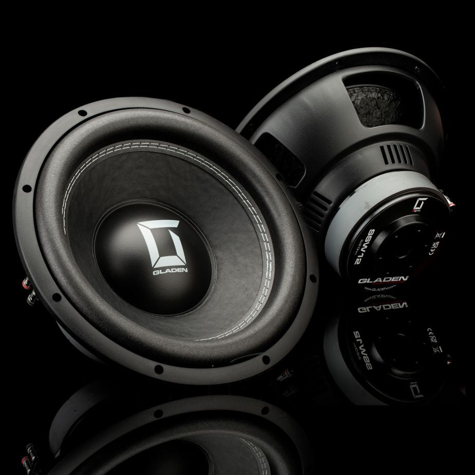 Dos subwoofers Gladen Sigma SSW12, uno orientado hacia delante y otro en ángulo, descansan sobre una superficie negra brillante, lo que realza los graves de este sistema de audio para coche de alta gama.
