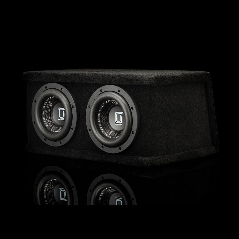 El Gladen Sigma SSWX065 Dual es un subwoofer rectangular negro con dos altavoces de 6,5, que aparece en una imagen sobre un fondo negro con su reflejo debajo.