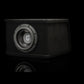 Un subwoofer Gladen Sigma SSWX08VB de 8 de color negro, con una caja rectangular, se encuentra sobre una superficie reflectante; en el centro de la membrana del altavoz se aprecia un logotipo plateado.