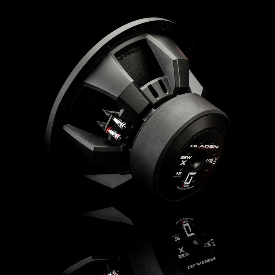 Un subwoofer Gladen Sigma SSWx12, con i cavi a vista, è posizionato di traverso su una superficie riflettente, mettendo in risalto la raffinata lavorazione artigianale del suo corpo nero.