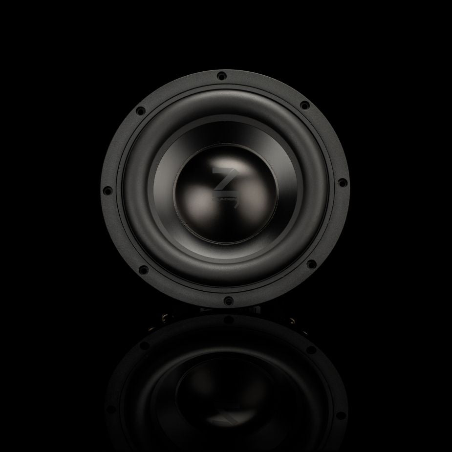 Un subwoofer Gladen Zeta 08, redondo y negro, ocupa el centro de una superficie brillante, mientras que una iluminación tenue resalta sus graves precisos y su elegante acabado.