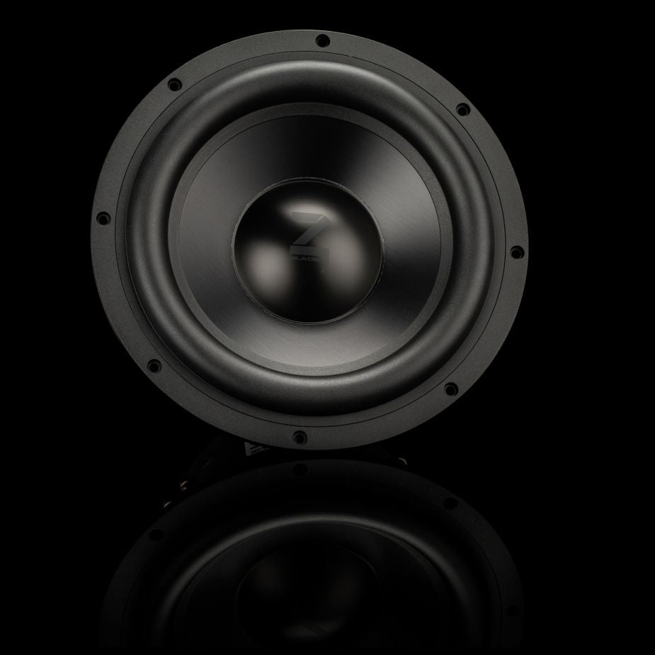 Se muestra un subwoofer Gladen Zeta 10 en color negro con el centro brillante sobre un fondo oscuro, lo que resalta los potentes graves y deja entrever un sutil reflejo en la parte inferior.