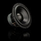 Un subwoofer Gladen Zeta 10 de color negro está colocado en ángulo sobre una superficie reflectante, dejando a la vista su cableado y los orificios de montaje, lo que lo hace ideal para una reproducción de graves profundos en tu equipo.